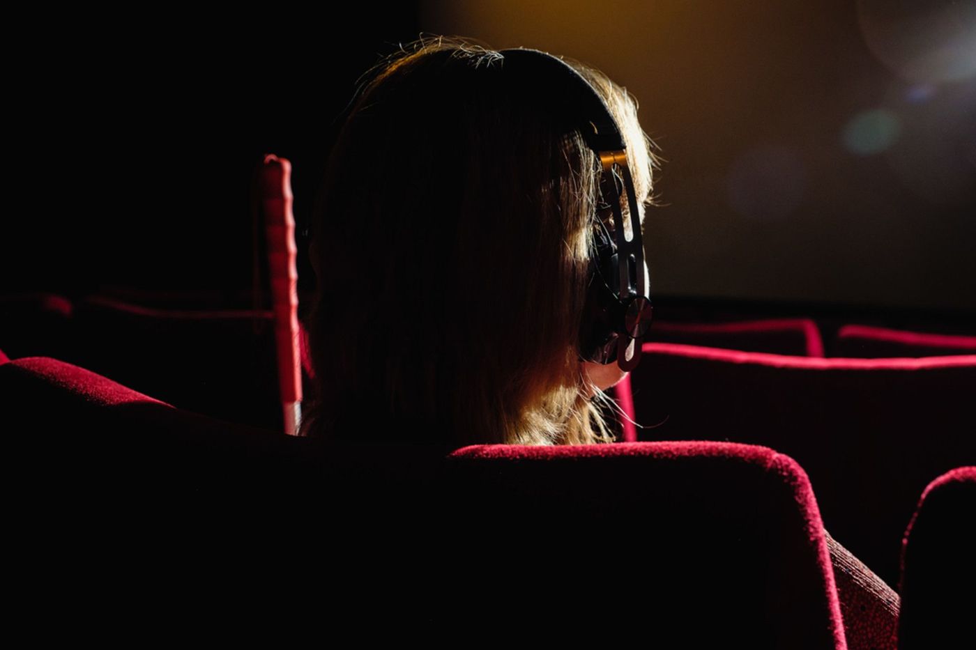 Das Foto zeigt eine Person von hinten, die im roten Kinosessel sitzt. Sie trägt einen Kopfhörer. Neben ihr ist ein Stab aufgestellt.